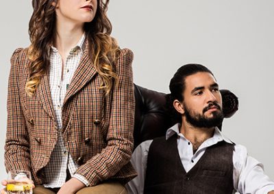 Female model in tweed jacket sits on am of chair holding drink whilst man sits and looks into the distance